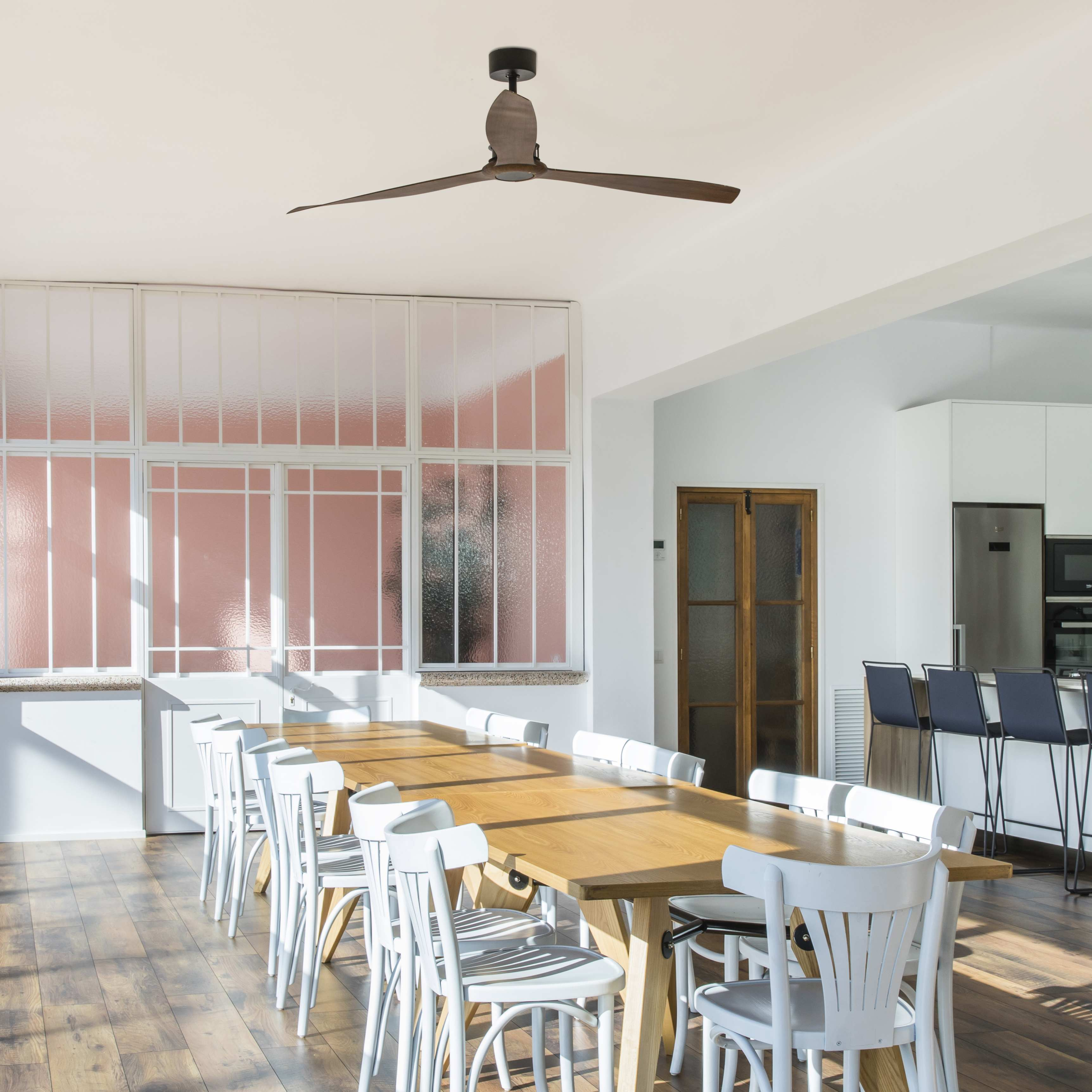 Sala da pranzo con un lungo tavolo di legno, sedie bianche e un ventilatore a soffitto. Grandi finestre, interni luminosi.