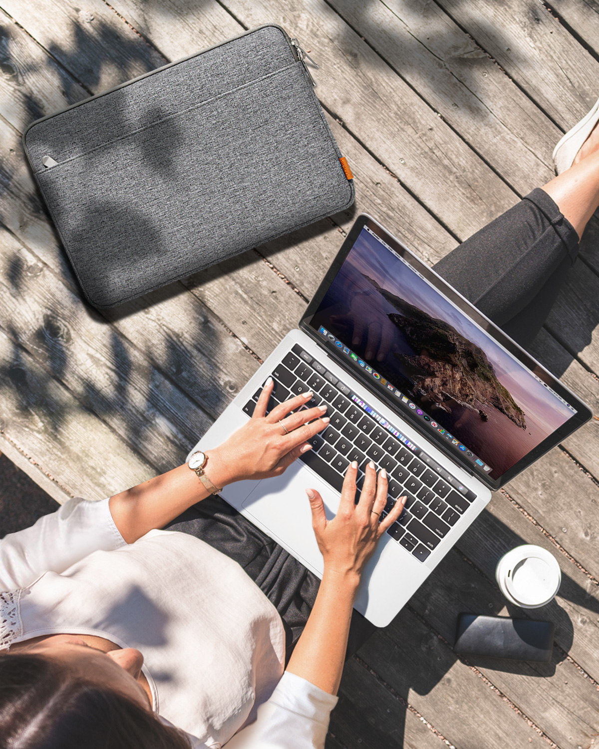 Frau benutzt Laptop, auf Holzplanken sitzend. Graue Laptoptasche und Kaffeetasse daneben.