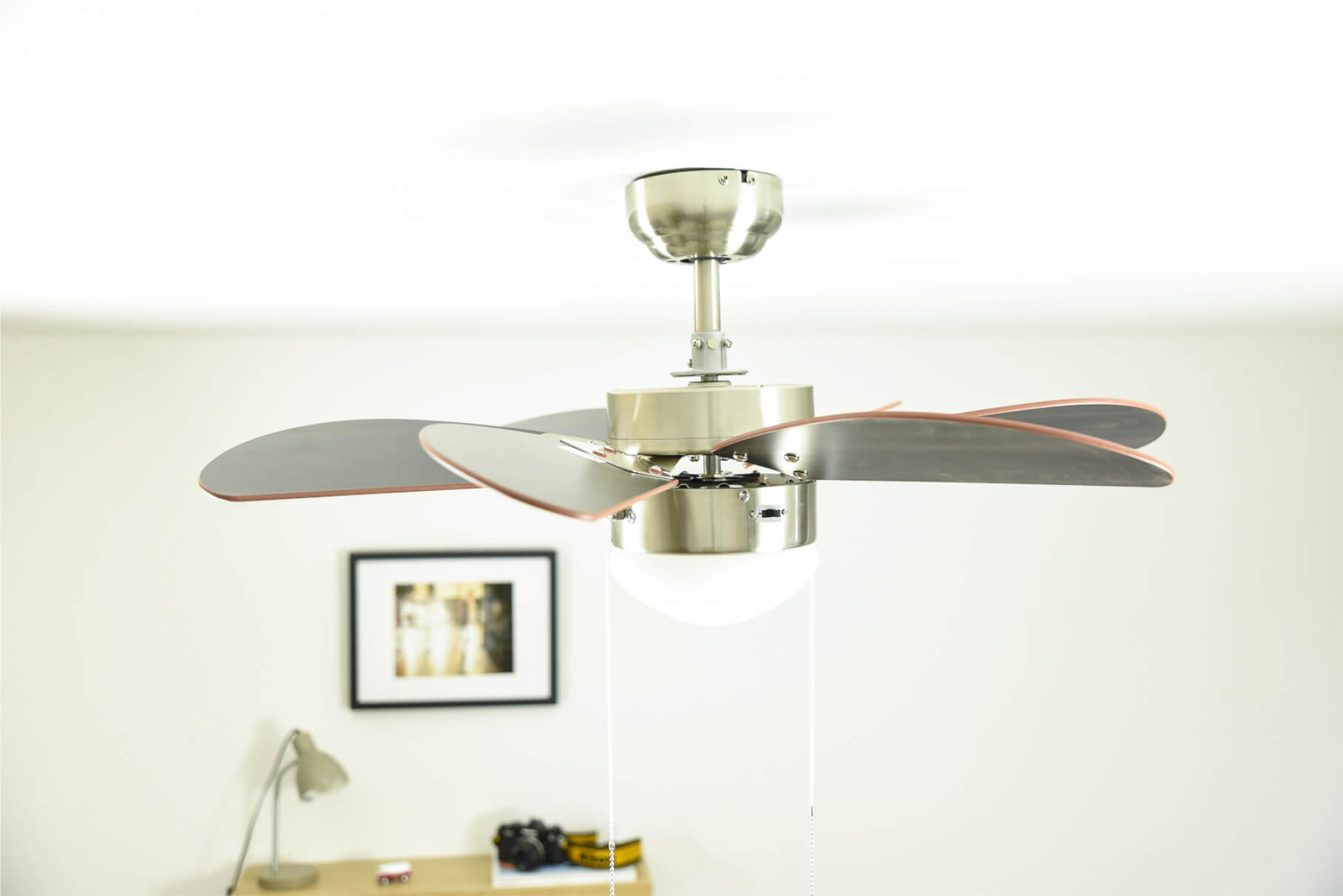 Ein Deckenventilator mit silbernen Flügeln und einer Leuchte hängt in einem Raum. Ein gerahmtes Bild und eine Lampe sind im Hintergrund.