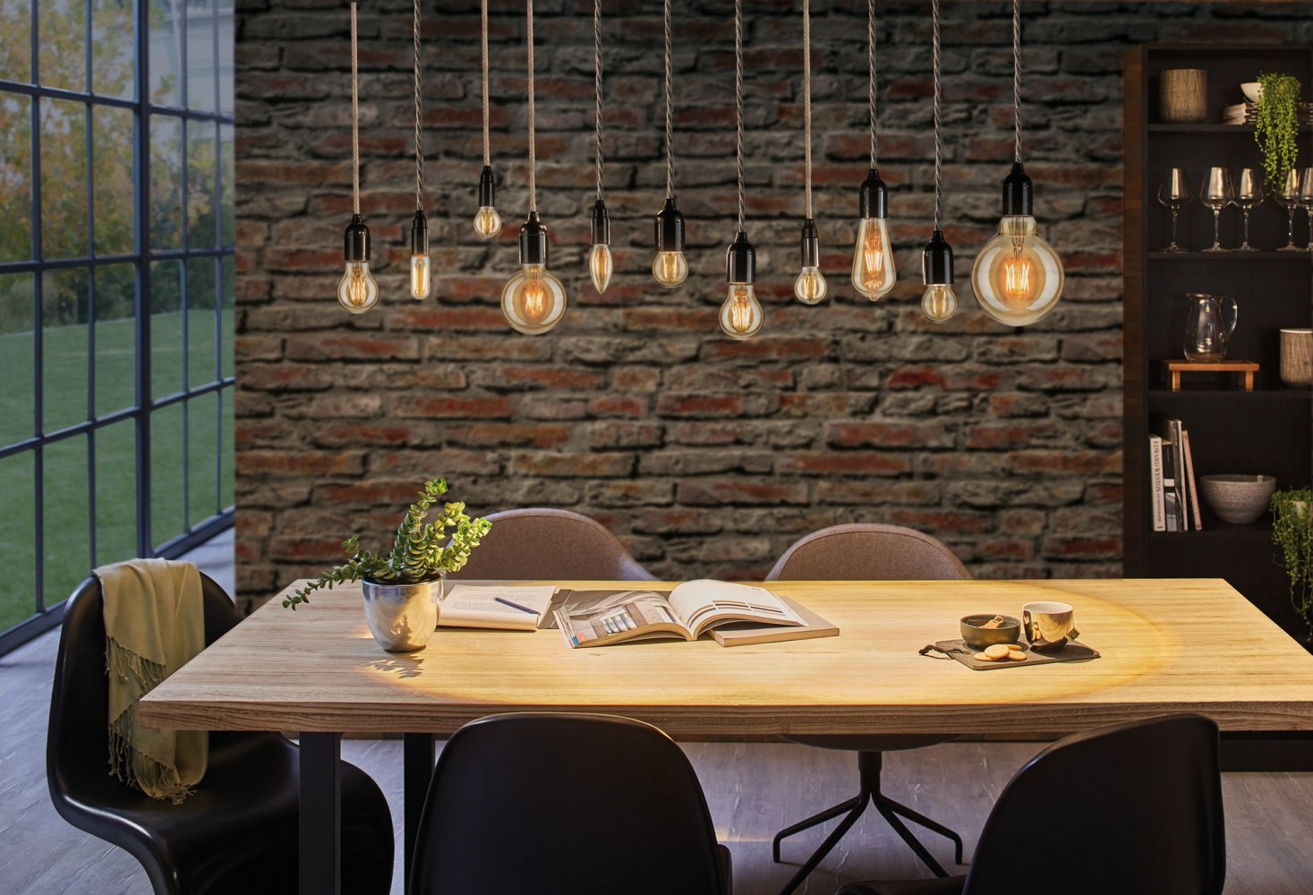 Jadalnia z drewnianym stołem i wiszącymi żarówkami. W tle ceglana ściana.