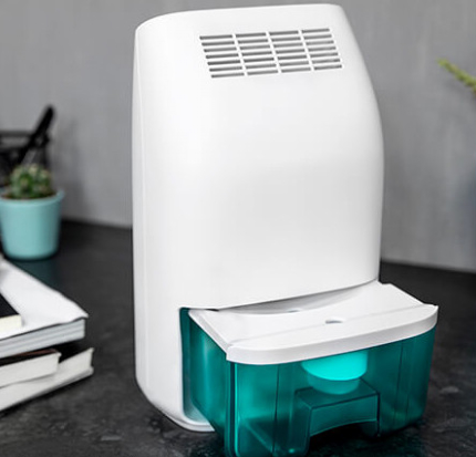 Biały osuszacz powietrza z turkusowym zbiornikiem na wodę. Stoi na ciemnej powierzchni.