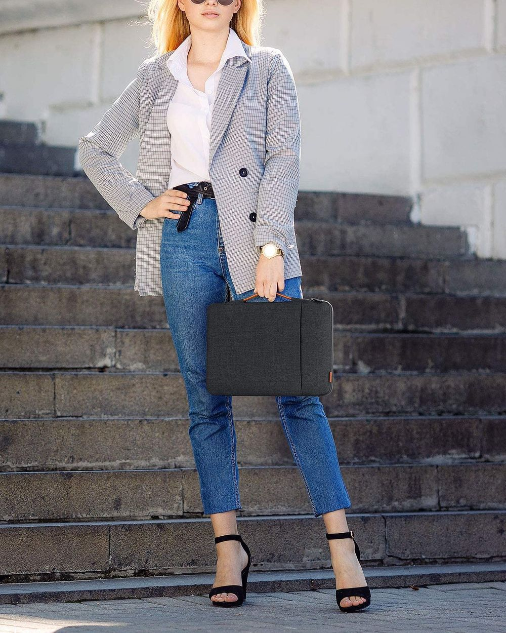 Frau in Blazer, Jeans und Absätzen hält eine Tasche. Treppen im Hintergrund. Sonniger Tag.