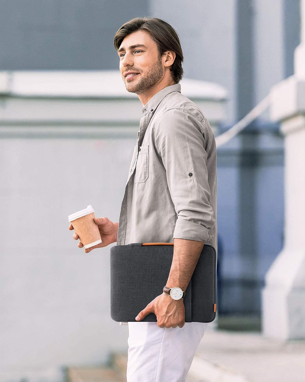 Ein Mann in hellem Hemd hält eine Kaffeetasse und eine Laptoptasche, geht draußen.