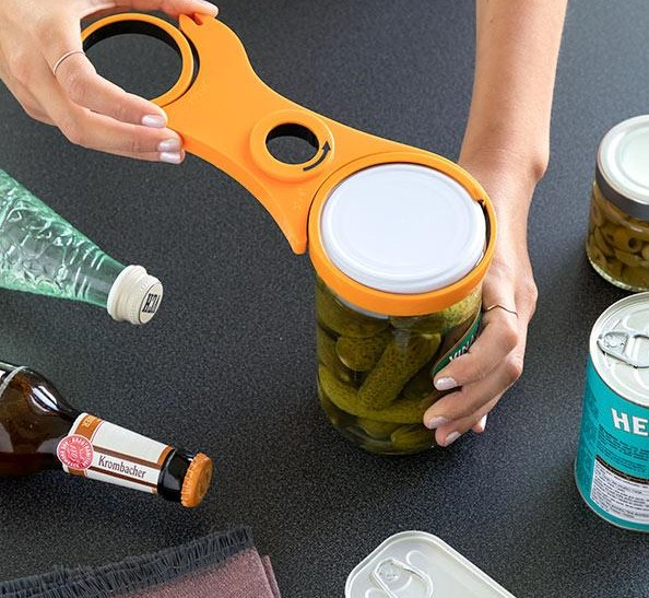 Person öffnet ein Glas mit einem orangefarbenen Glasöffner. Weitere Artikel: Bierflasche und Konserven.