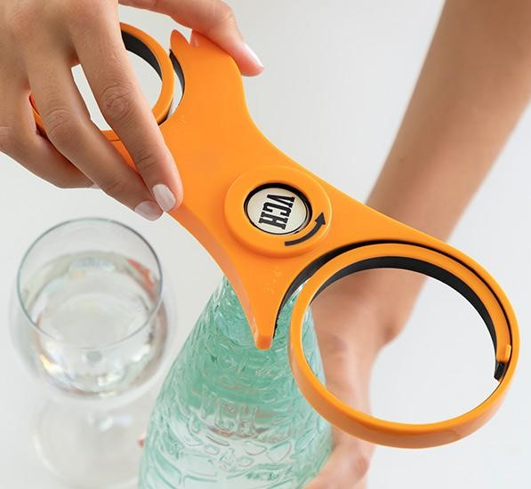Ein orangefarbener Flaschenöffner wird von zwei Händen gehalten, eine Flasche im Fokus.
