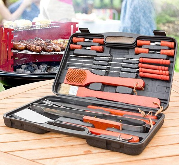 Geöffnete schwarze Aktentasche mit Grillwerkzeug, roter Grill im Hintergrund, Holzoberfläche.