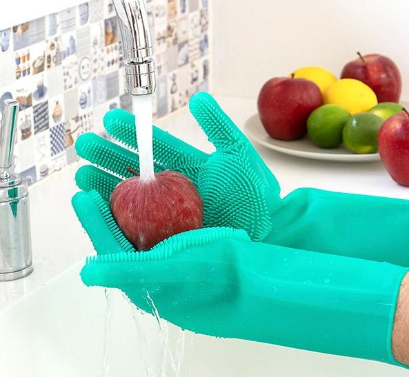 Grüne Handschuhe waschen einen roten Apfel unter einem Wasserhahn. Hintergrund: Früchte auf einem Teller, weißes Waschbecken.