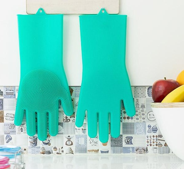Zwei grüne, handförmige Silikonhandschuhe hängen an einer gefliesten Wand. Hintergrund zeigt Früchte.
