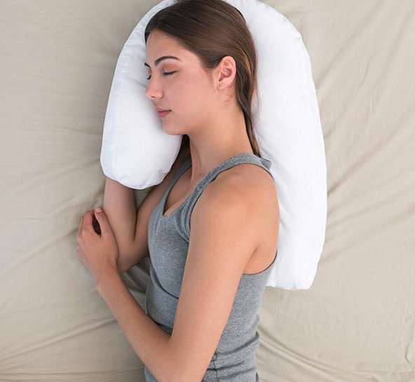Frau schläft im Bett mit U-förmigem weißen Kissen, trägt graues Tanktop.