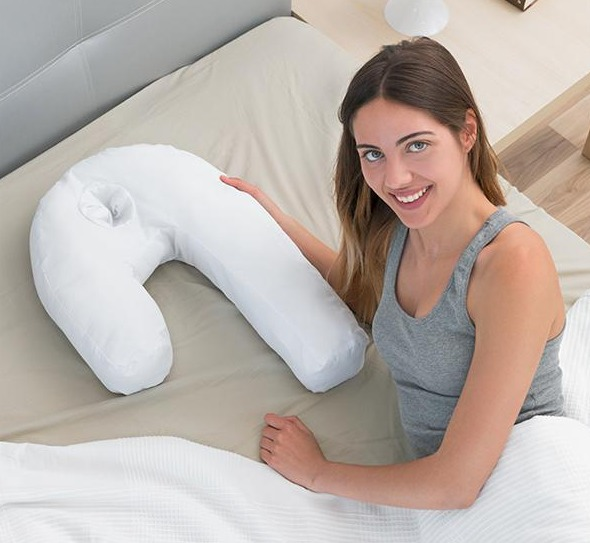 Frau in einem Bett, lächelnd, hält ein weißes U-förmiges Kissen. Beige Bettwäsche.