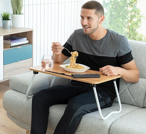 Un uomo seduto su un divano mangia spaghetti da un piatto su un tavolino portatile.