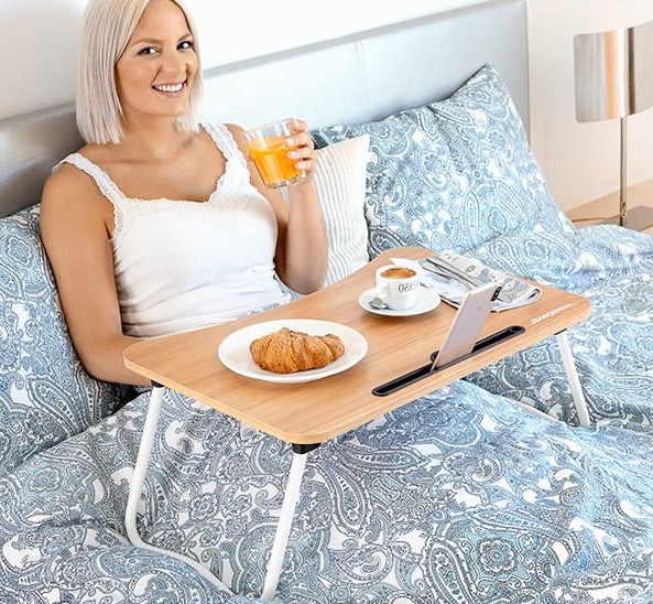 Donna a letto con laptop, caffè e cibo su vassoio in legno. Tiene bicchiere di succo.