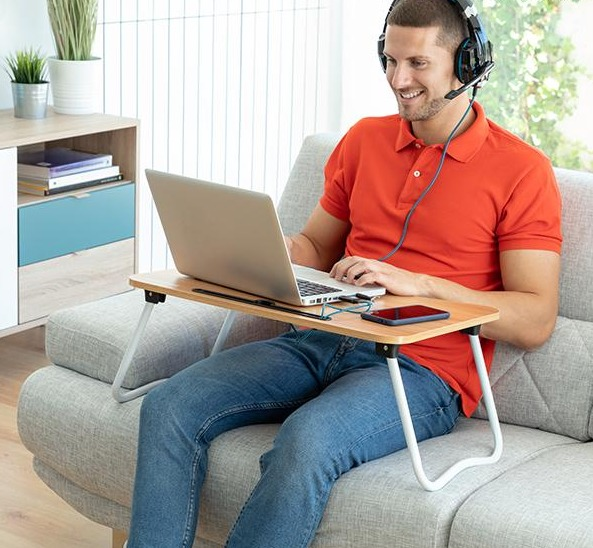 Uomo con cuffie e laptop seduto su un divano. Una scrivania per laptop è sulle sue ginocchia.
