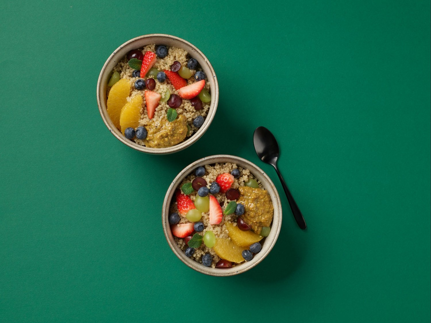 Zwei Schalen mit Obst- und Quinoasalat auf grüner Oberfläche. Löffel rechts. Draufsicht.