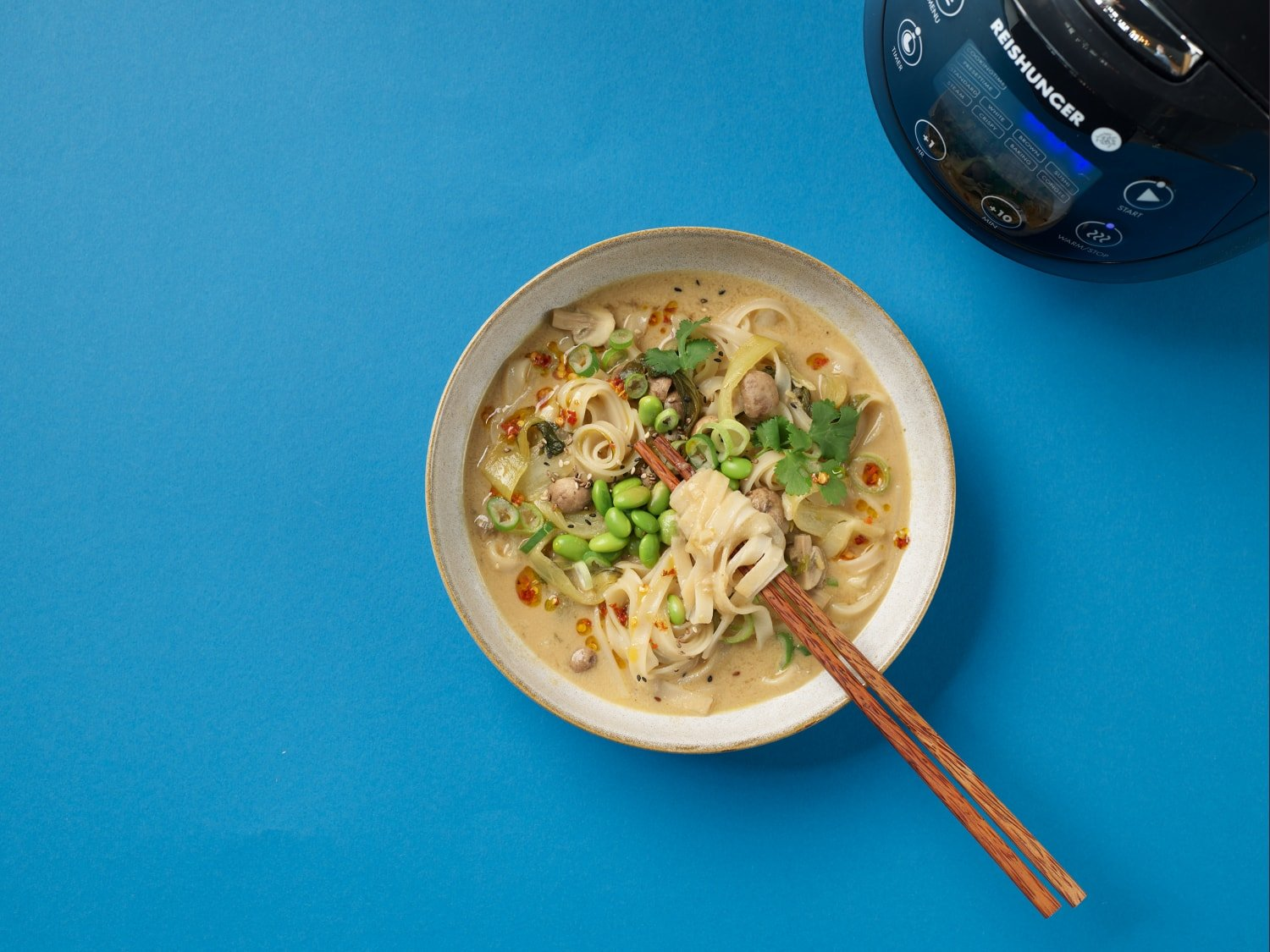 Schüssel Suppe mit Essstäbchen auf blauem Untergrund, Reiskocher teilweise sichtbar.
