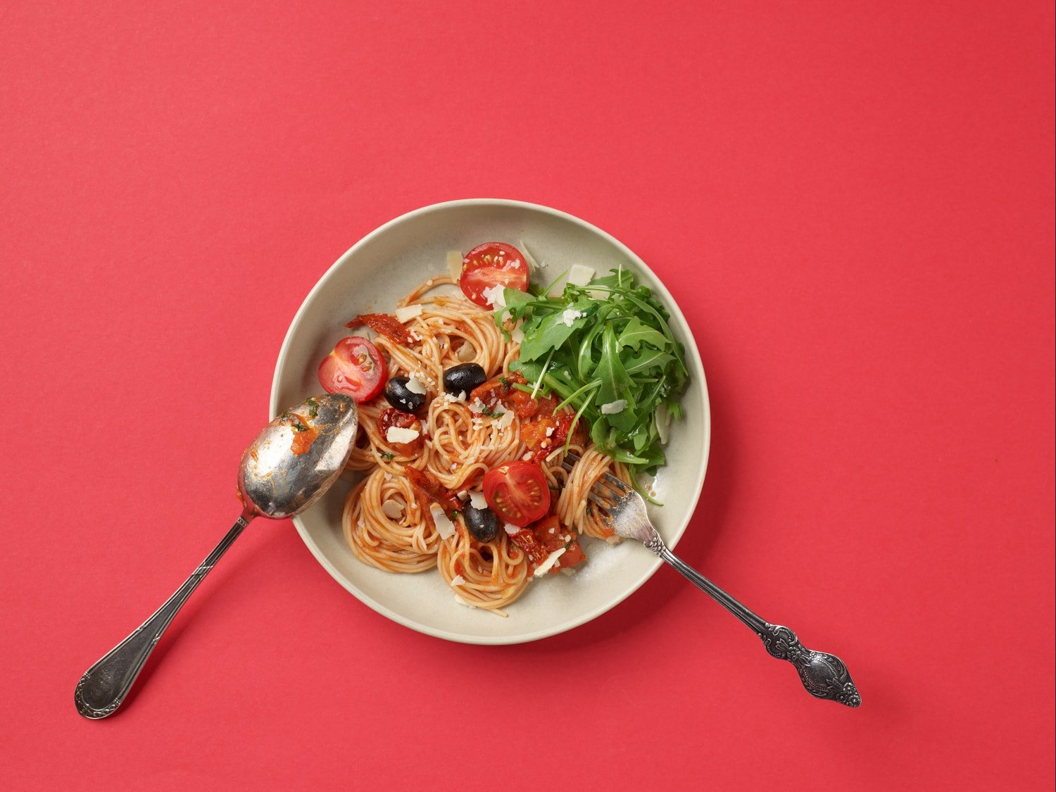 Spaghetti mit Tomaten, Oliven und Rucola in einer Schüssel mit Löffel und Gabel.