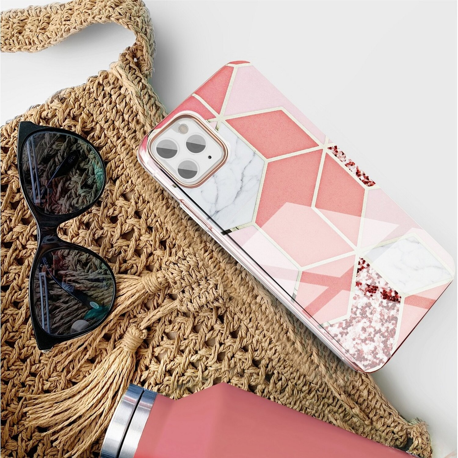 Handyhülle mit rosa geometrischem Design, Sonnenbrille und rosa Trinkflasche auf einer gewebten Tasche.