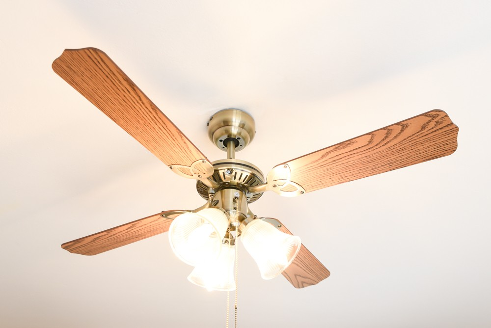Ventilatore a soffitto con quattro pale in legno e luci. Fissaggio centrale color oro. Soffitto bianco.