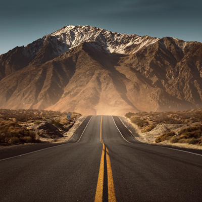 Ansicht einer Straße, die in eine Wüstenlandschaft mit einem Berg im Hintergrund führt.