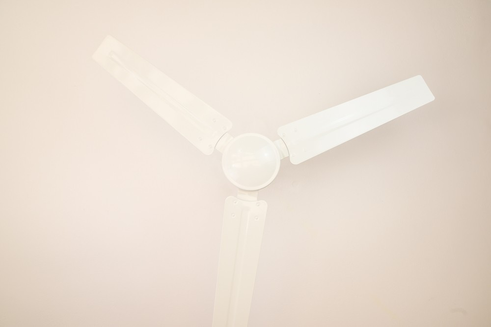 Ein weißer Deckenventilator mit drei Flügeln vor einer weißen Decke. Die Perspektive ist nach oben gerichtet.
