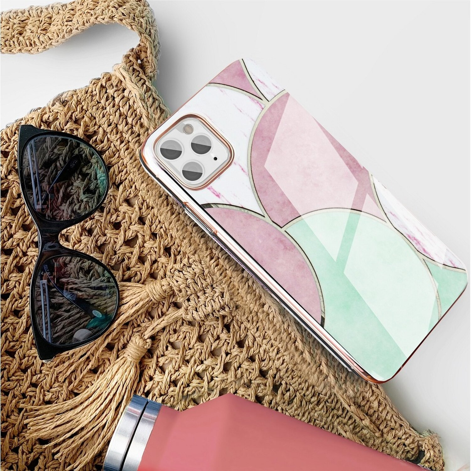 Handyhülle auf einer Tasche, Sonnenbrille und Wasserflasche. Das Etui hat rosa und grüne Formen.