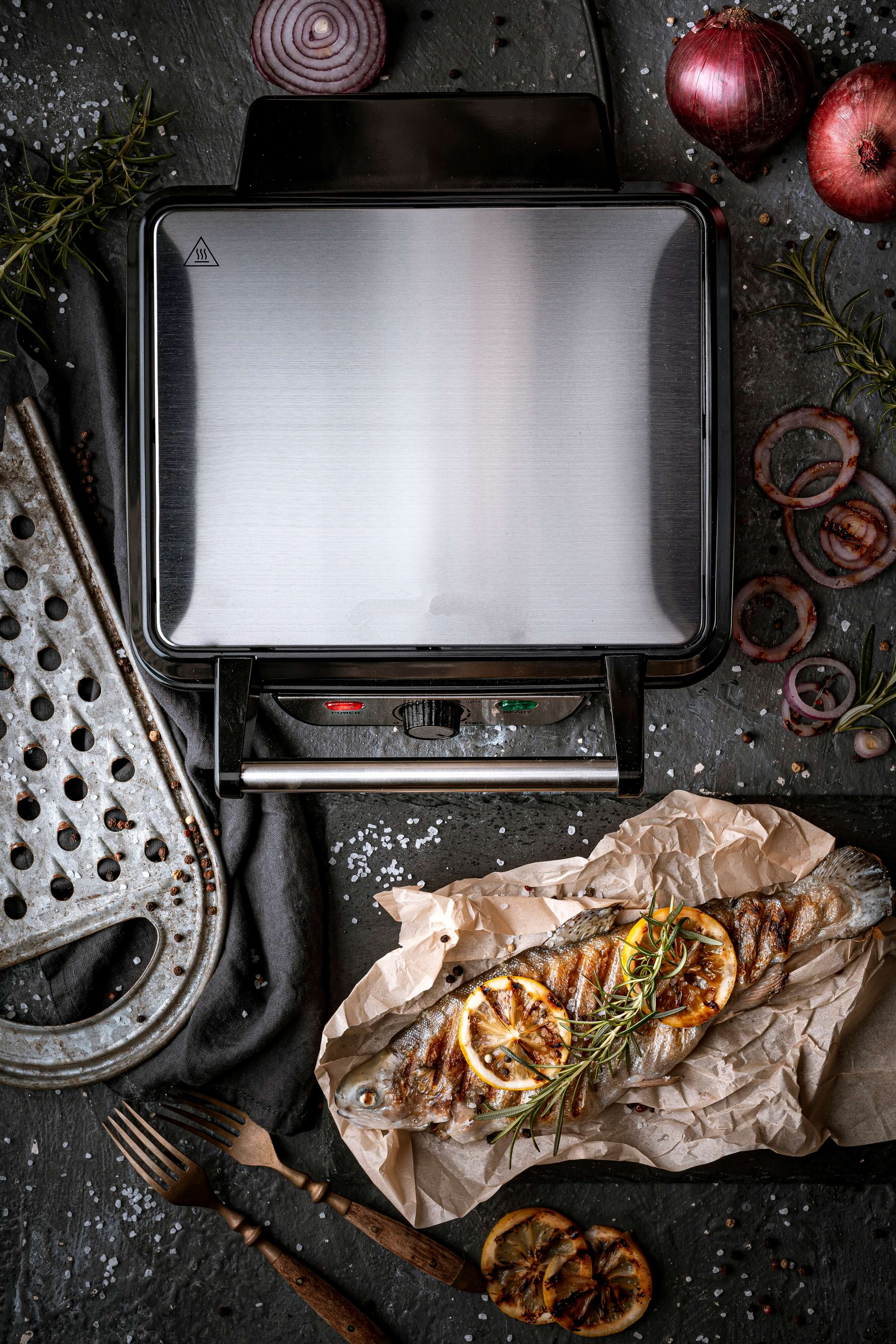 Draufsicht auf einen Grill mit gegrilltem Fisch, Zitronenscheiben und Rosmarin. Rote Zwiebeln und ein Reibeisen.