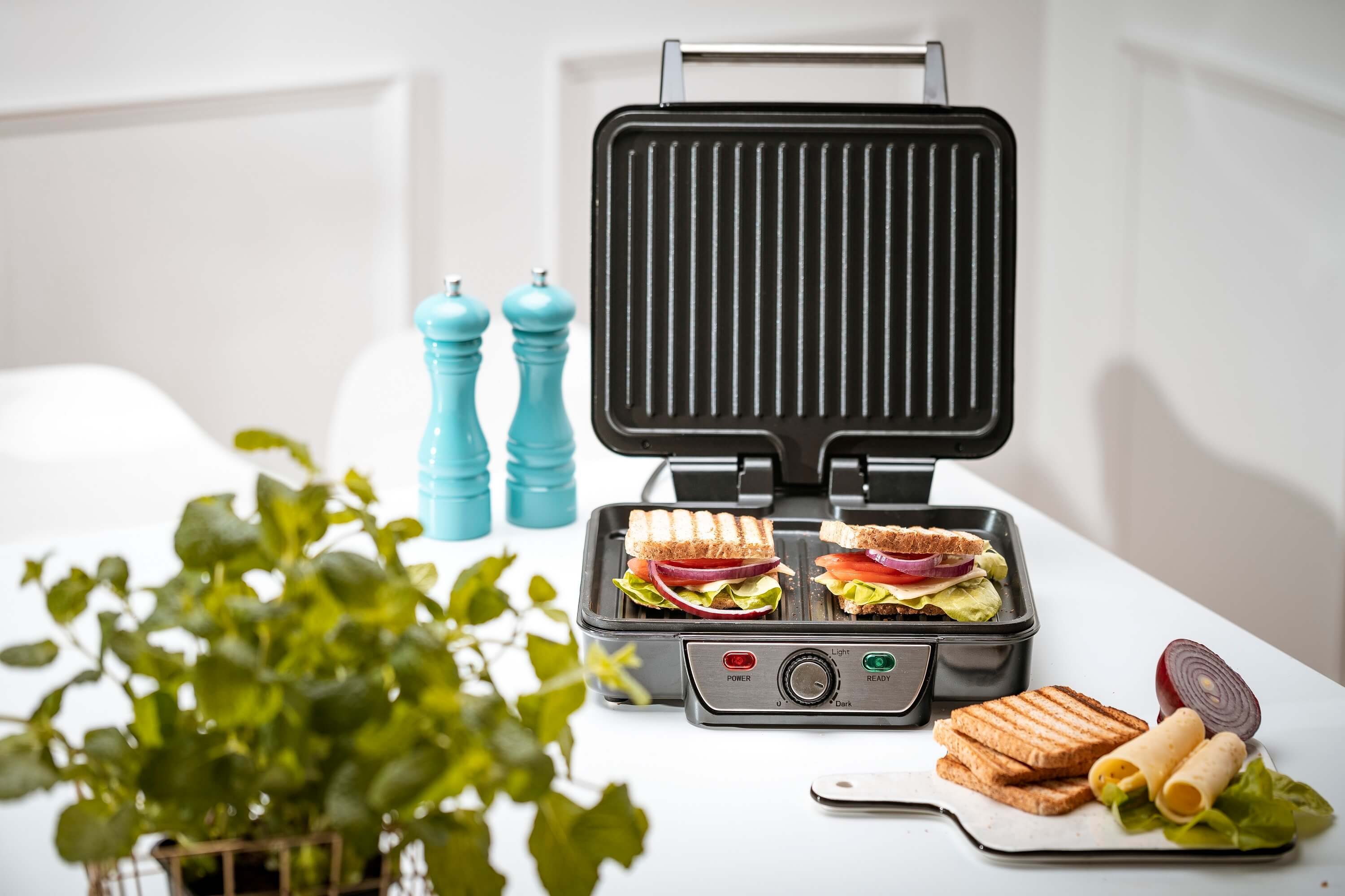 Ein Sandwichgrill mit zwei Sandwiches, Gemüse und Salz- und Pfefferstreuern auf einem Tisch.