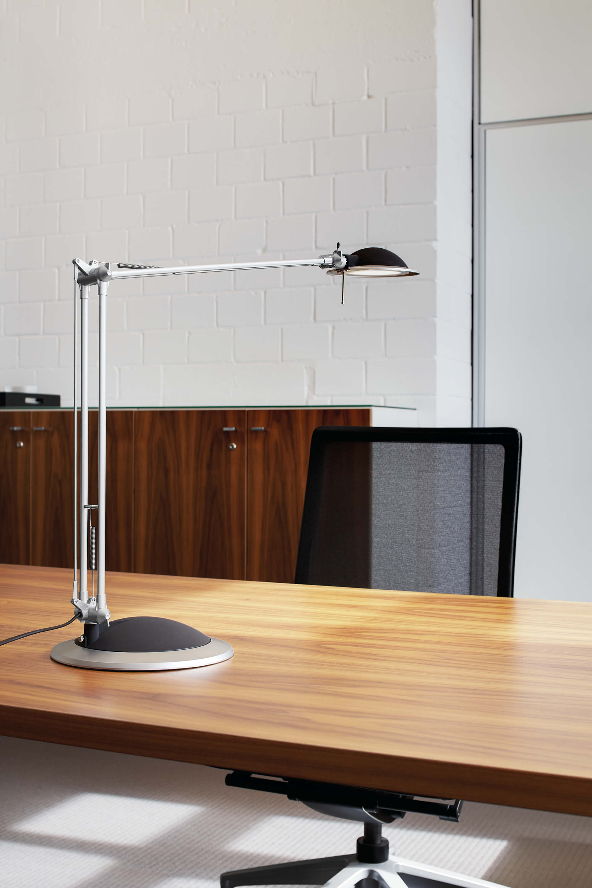 Lampa biurkowa z czarną podstawą i głowicą oraz srebrnym ramieniem stoi na drewnianym biurku.