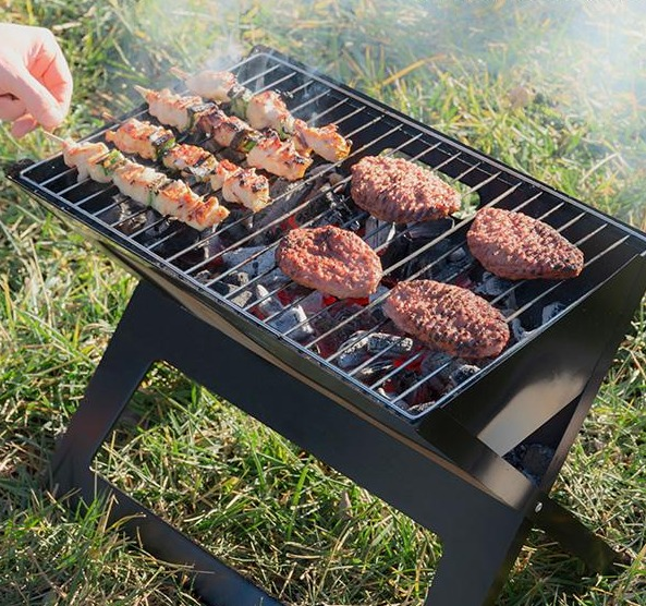 Griglia con cibo, tra cui kebab e hamburger, cotti su carboni ardenti sull'erba.
