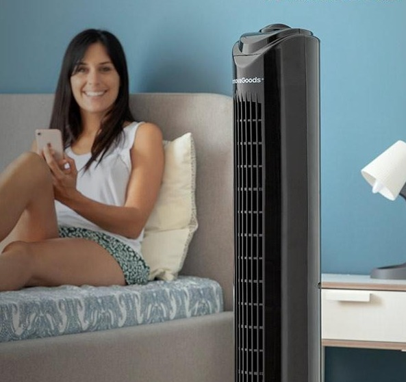 Ein schwarzer Turmventilator neben einer Frau, die mit einem Telefon auf einem Bett sitzt, mit einer Lampe auf einem Nachttisch.