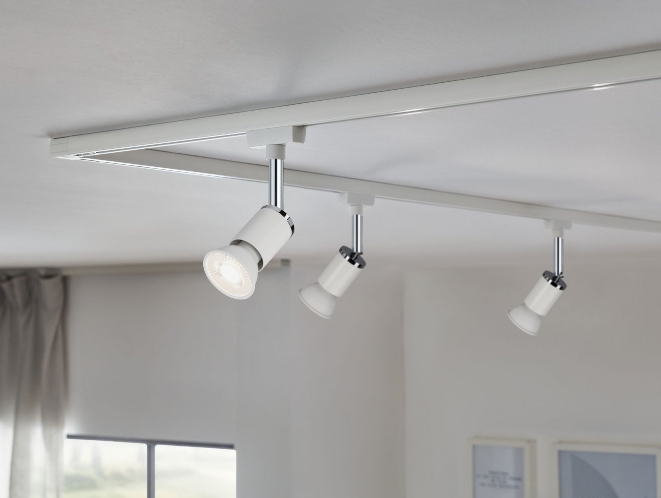 Trzy białe reflektory na białej szynie na białym suficie. Światła są skierowane w dół. Tłem jest biała ściana.