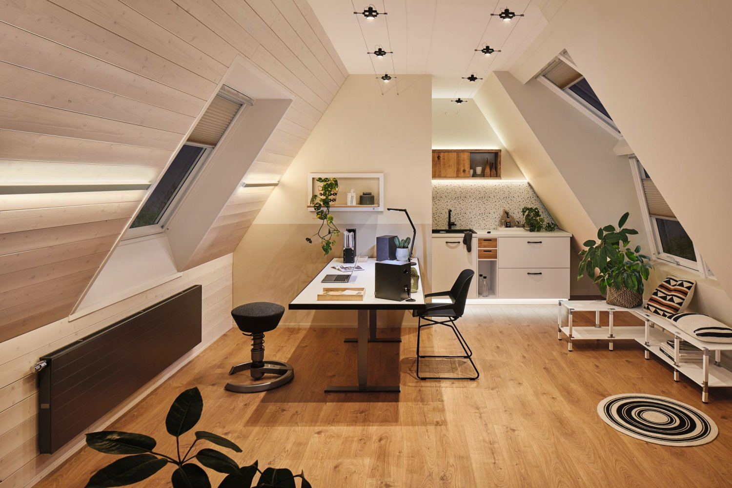 Biuro na poddaszu. Białe ściany, drewniana podłoga. Biurko, krzesło, rośliny i kuchnia.