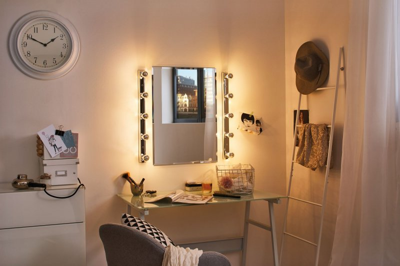 Una toeletta con specchio, luci e accessori, con un orologio e un cappello di lato.