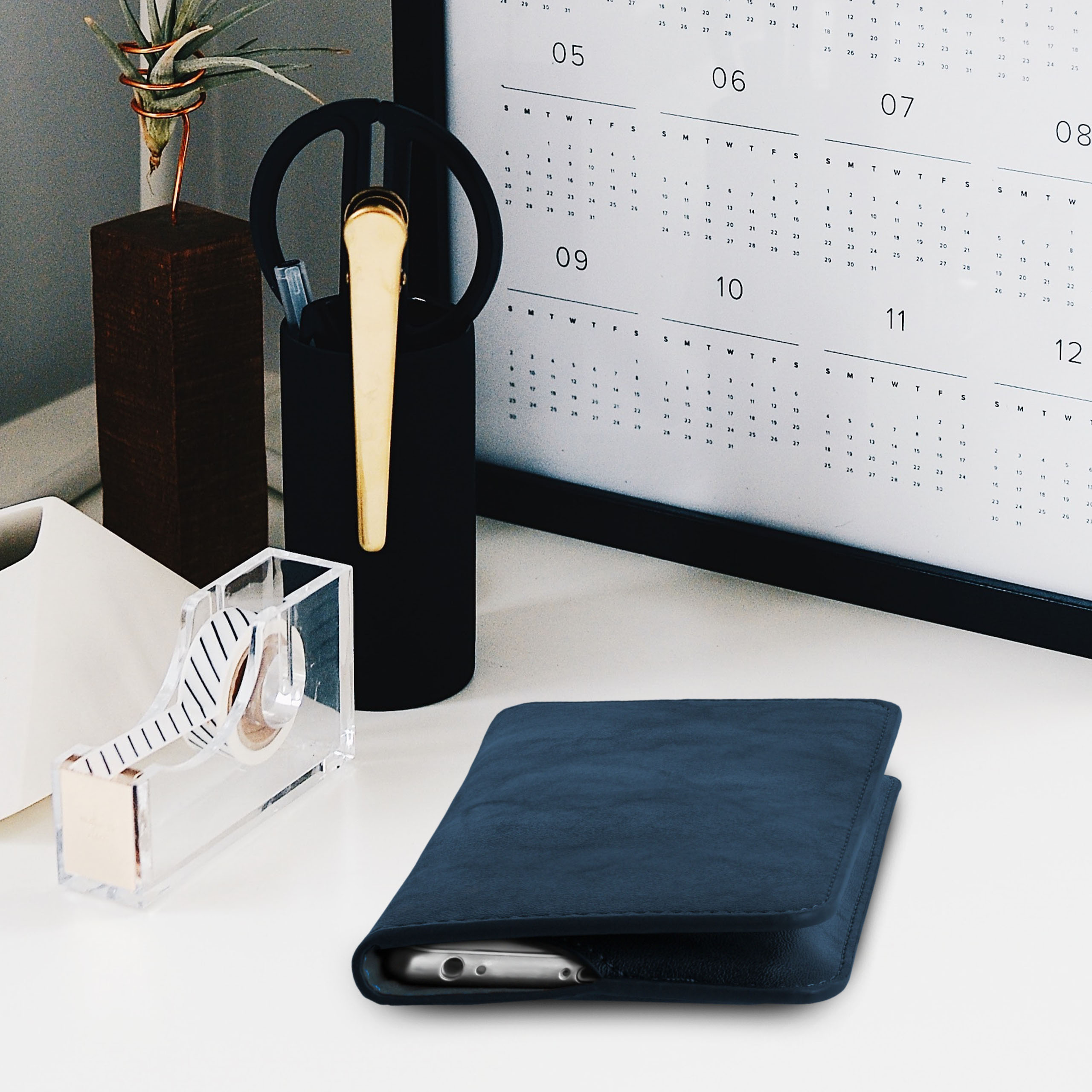 Schreibtisch-Setup mit Kalender, Stifthalter, Klebebandabroller und einer blauen Handytasche mit Telefon.