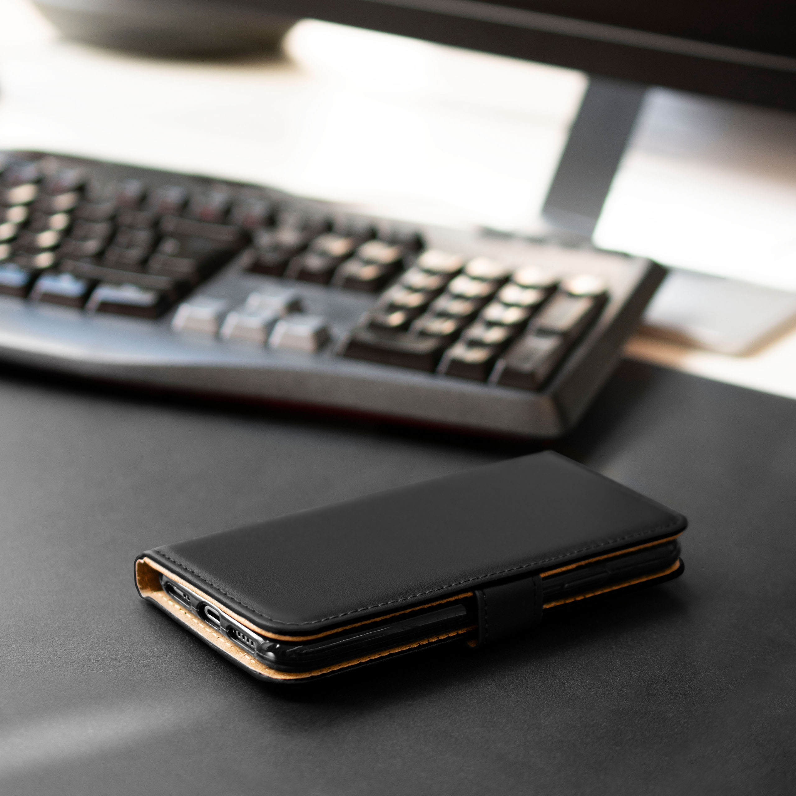 Schwarze Leder-Handyhülle mit Verschluss, auf schwarzem Schreibtisch, Tastatur unscharf im Hintergrund.
