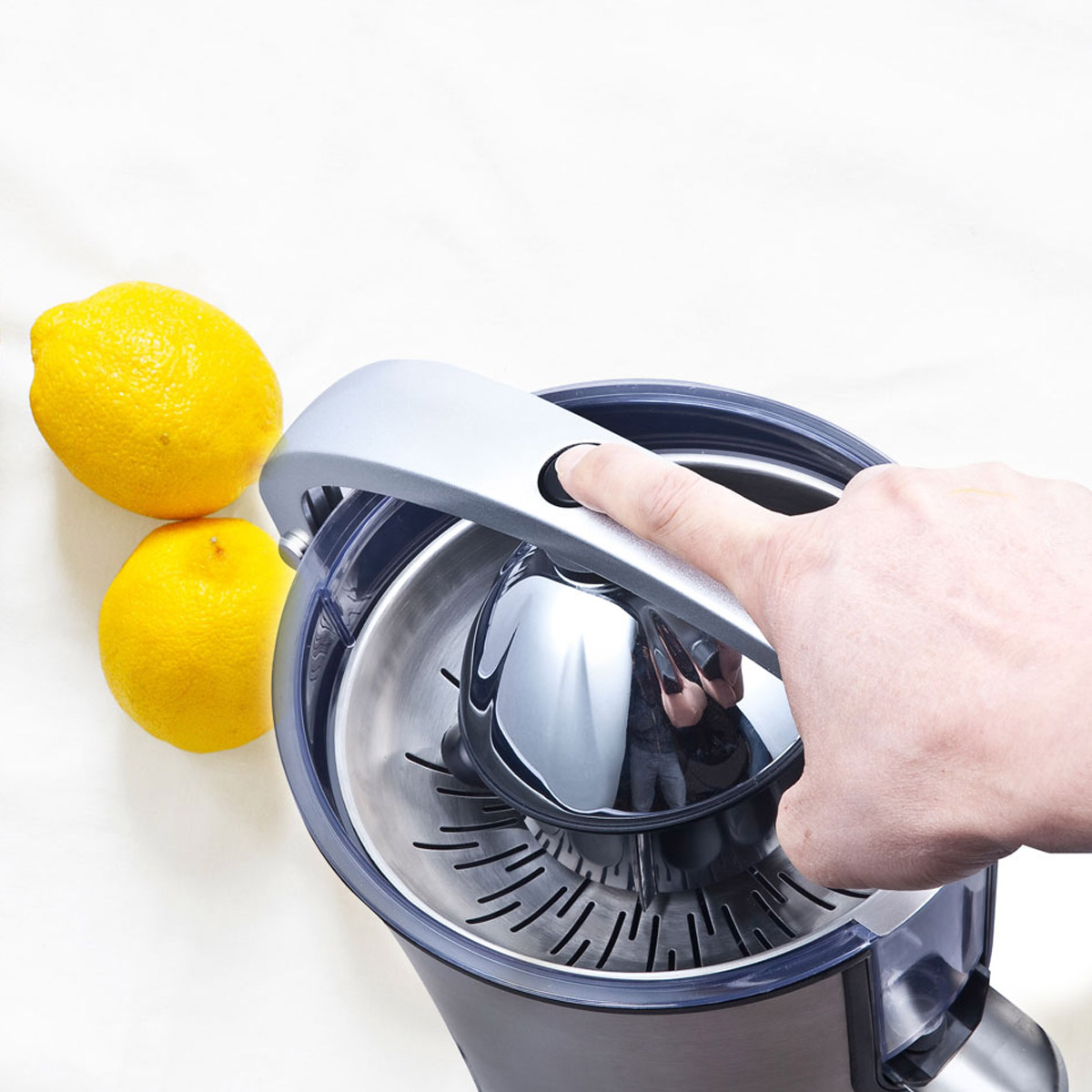 Ręka naciska przycisk na wyciskarce do soku. Dwie cytryny na białej powierzchni.