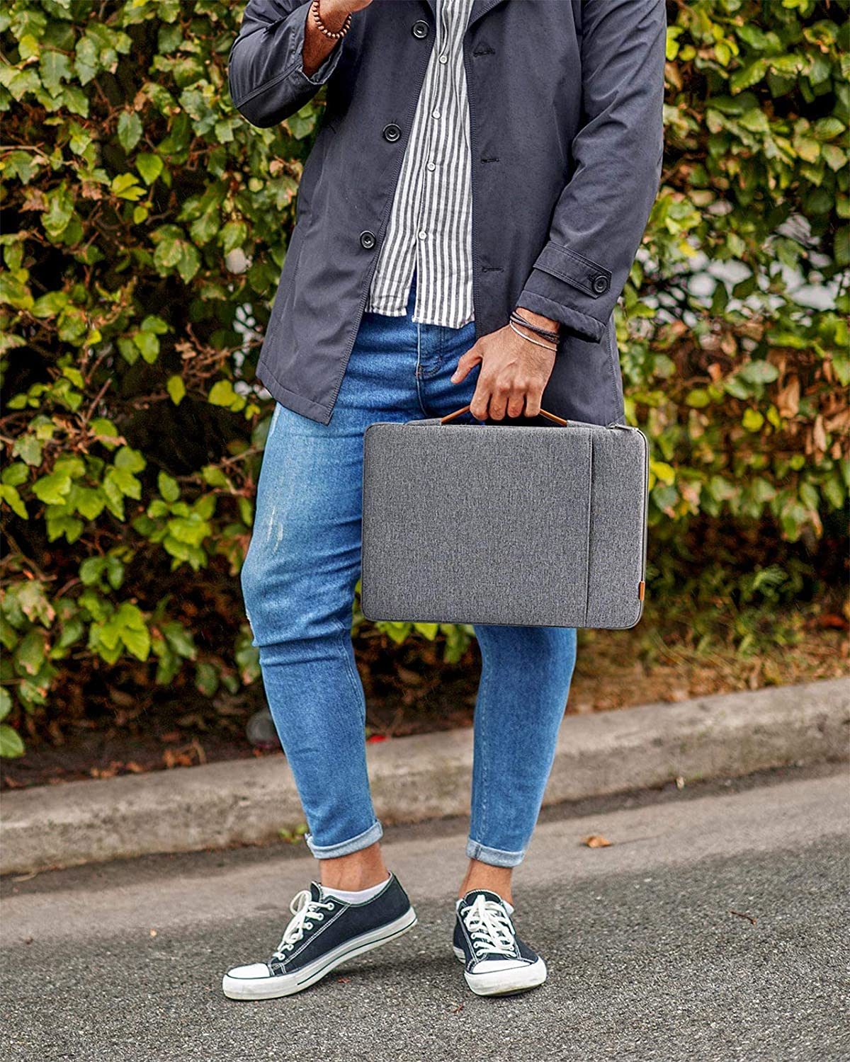 Ein Mann in Jeans und Jacke hält eine graue Laptoptasche. Er steht draußen auf dem Bürgersteig.