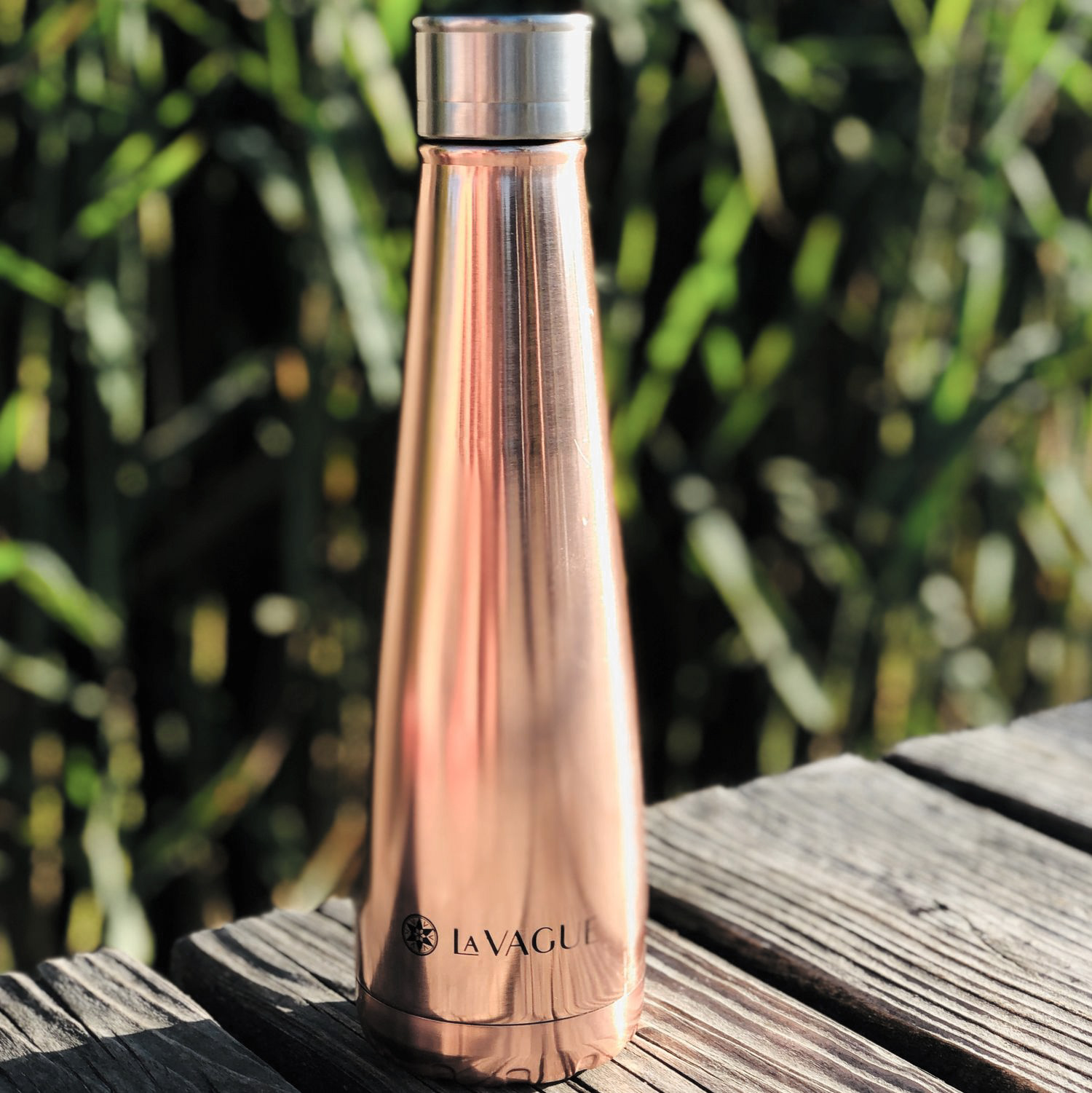 Botella de agua de oro rosa sobre una superficie de madera, fondo verde borroso, el logotipo 'LA VAGUE'.