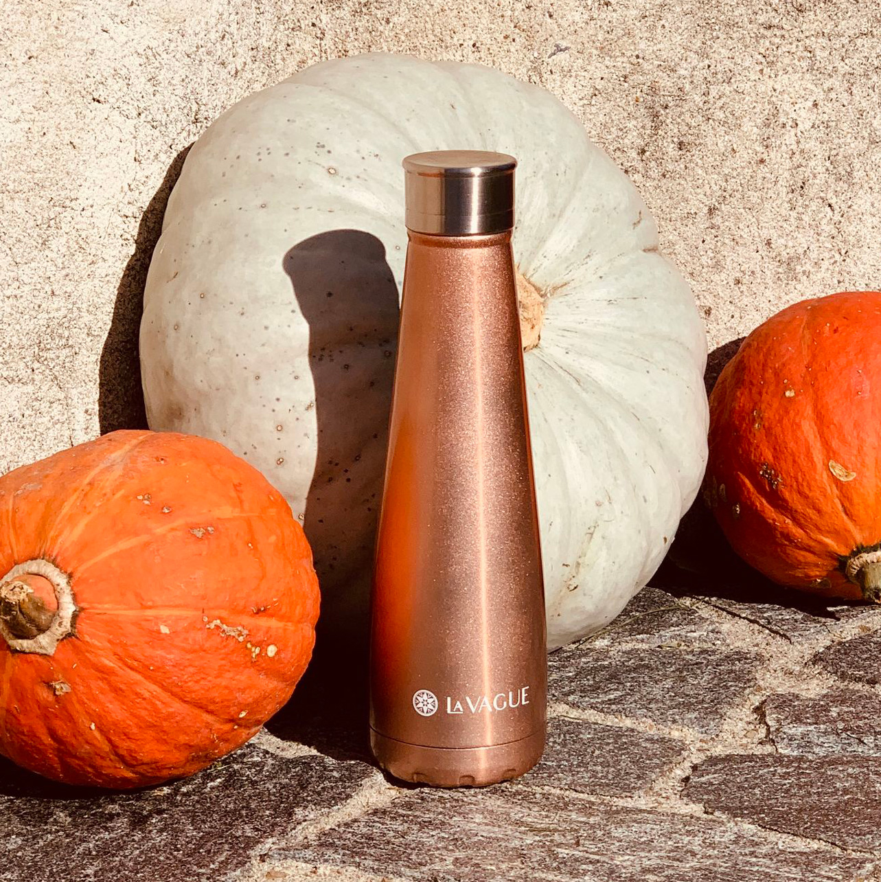 Una botella de agua de oro rosa frente a dos calabazas y una gran calabaza blanca sobre una superficie de piedra.