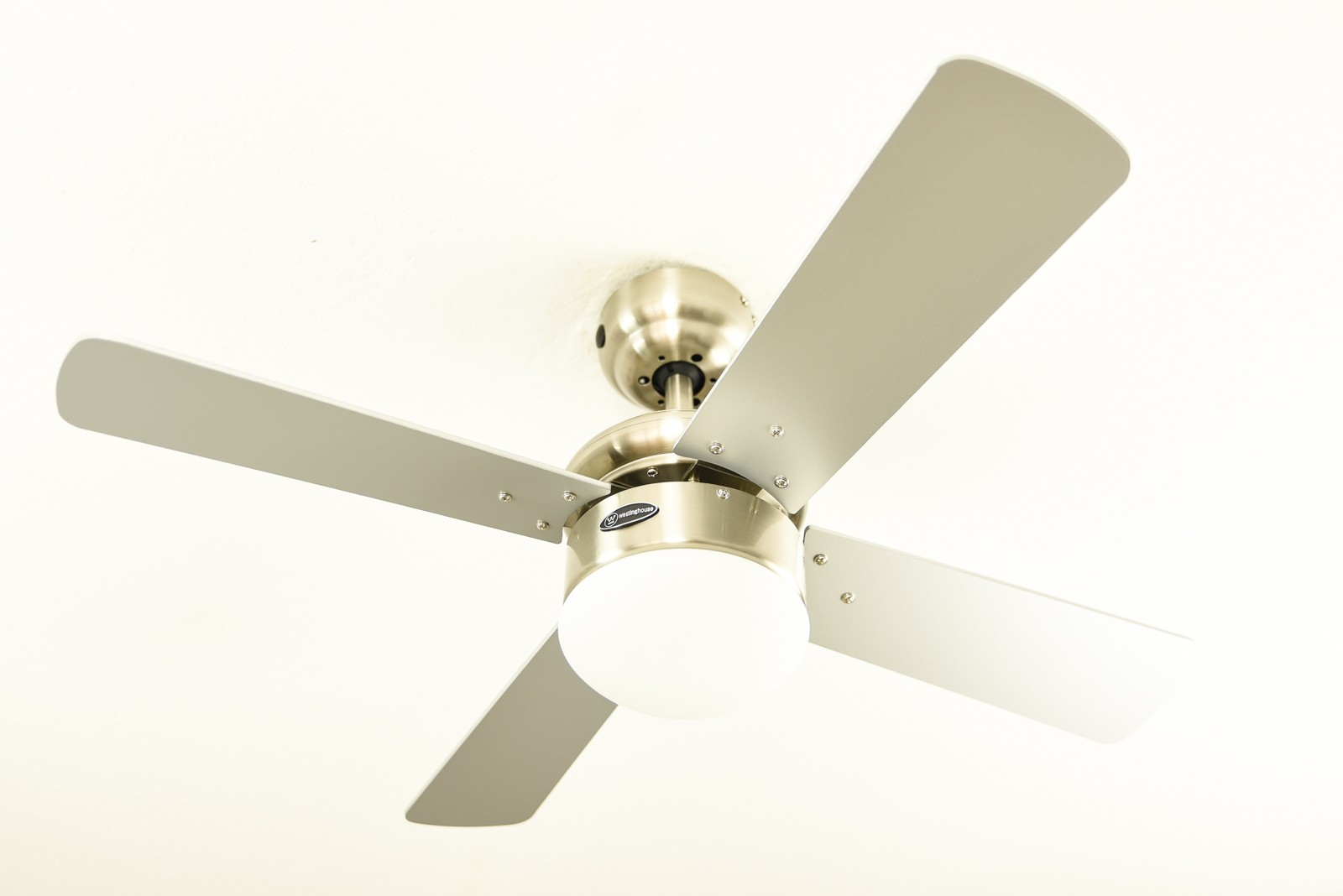 Ein Deckenventilator mit vier silbernen Flügeln und einem zentralen Licht. Der Ventilator befindet sich an einer weißen Decke.