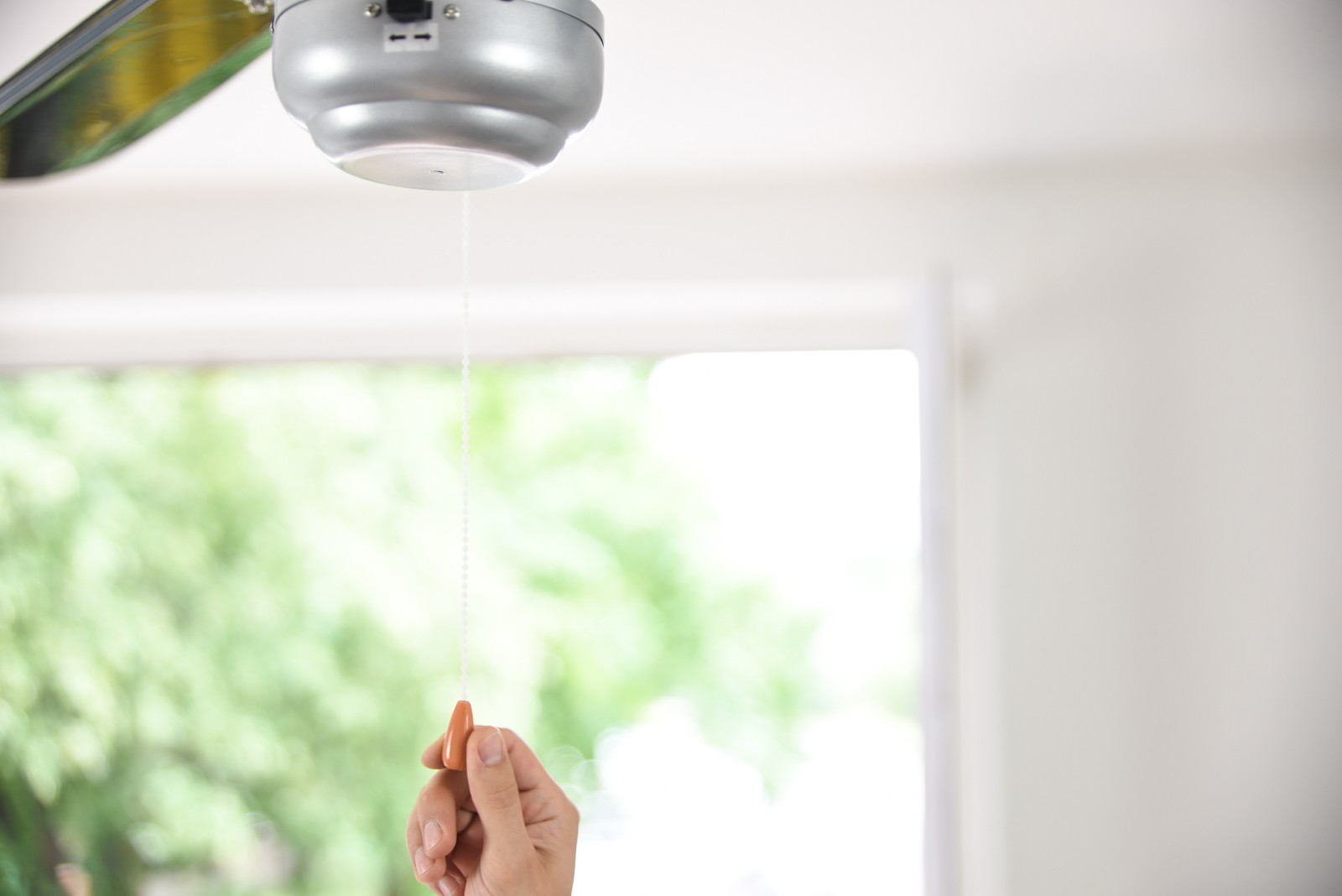 Una mano tira una corda per accendere un ventilatore da soffitto davanti a una finestra.