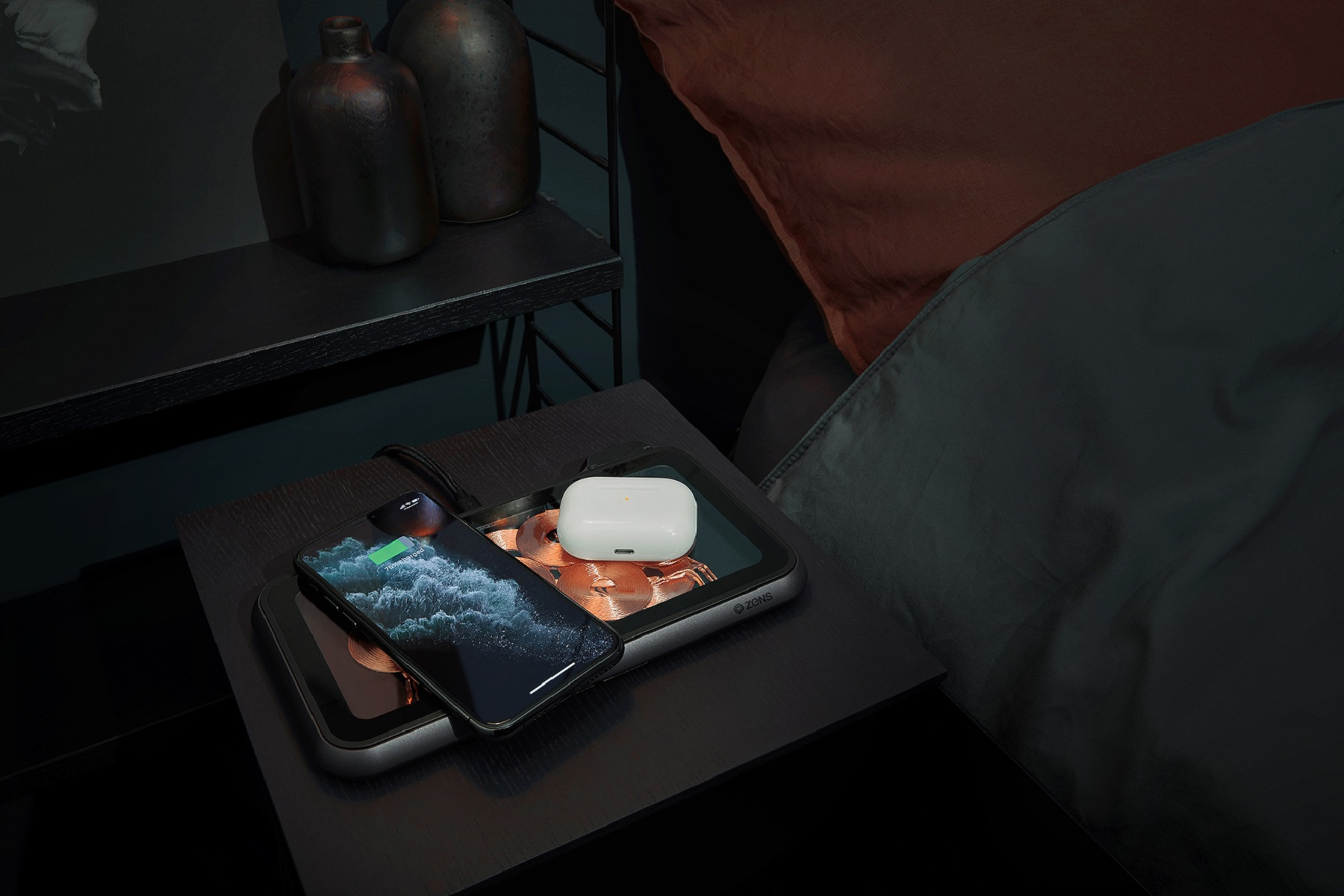 Ein Telefon und Ohrhörer laden auf einem Nachttisch neben einem Bett mit roter Decke.