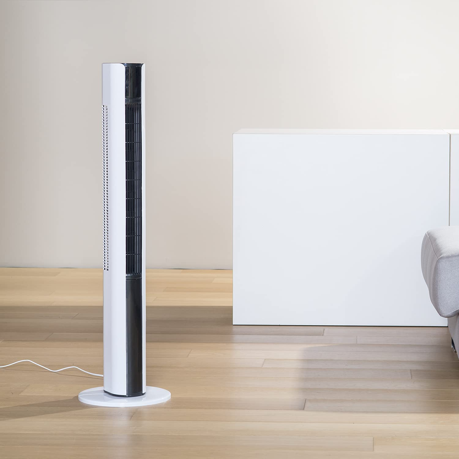 Biało-czarny wentylator stojący na drewnianej podłodze, z białą skrzynią w tle.