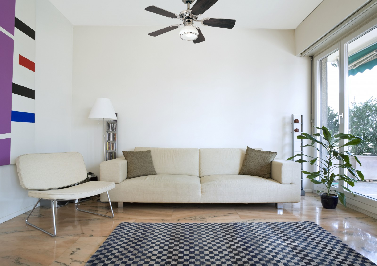Ein Wohnzimmer mit Sofa, Stuhl, Teppich und Deckenventilator. Weiße Wände und ein großes Fenster.
