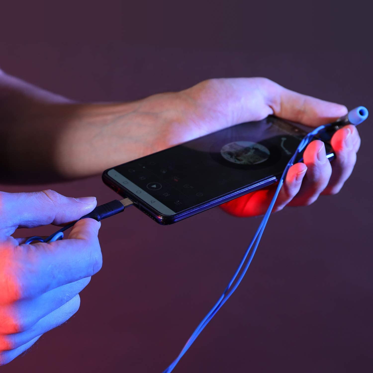 Persona che tiene in mano uno smartphone con un jack per cuffie collegato, illuminazione blu e rossa.