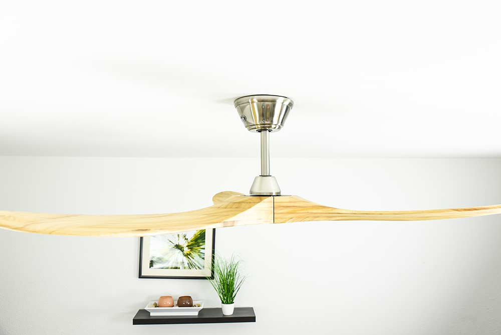 Un ventilatore da soffitto in legno con pale e un motore argentato. Una piccola immagine su un muro bianco.