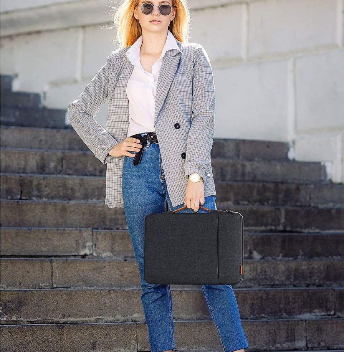 Mujer con gafas de sol, jeans y blazer, llevando una funda de portátil gris oscuro.