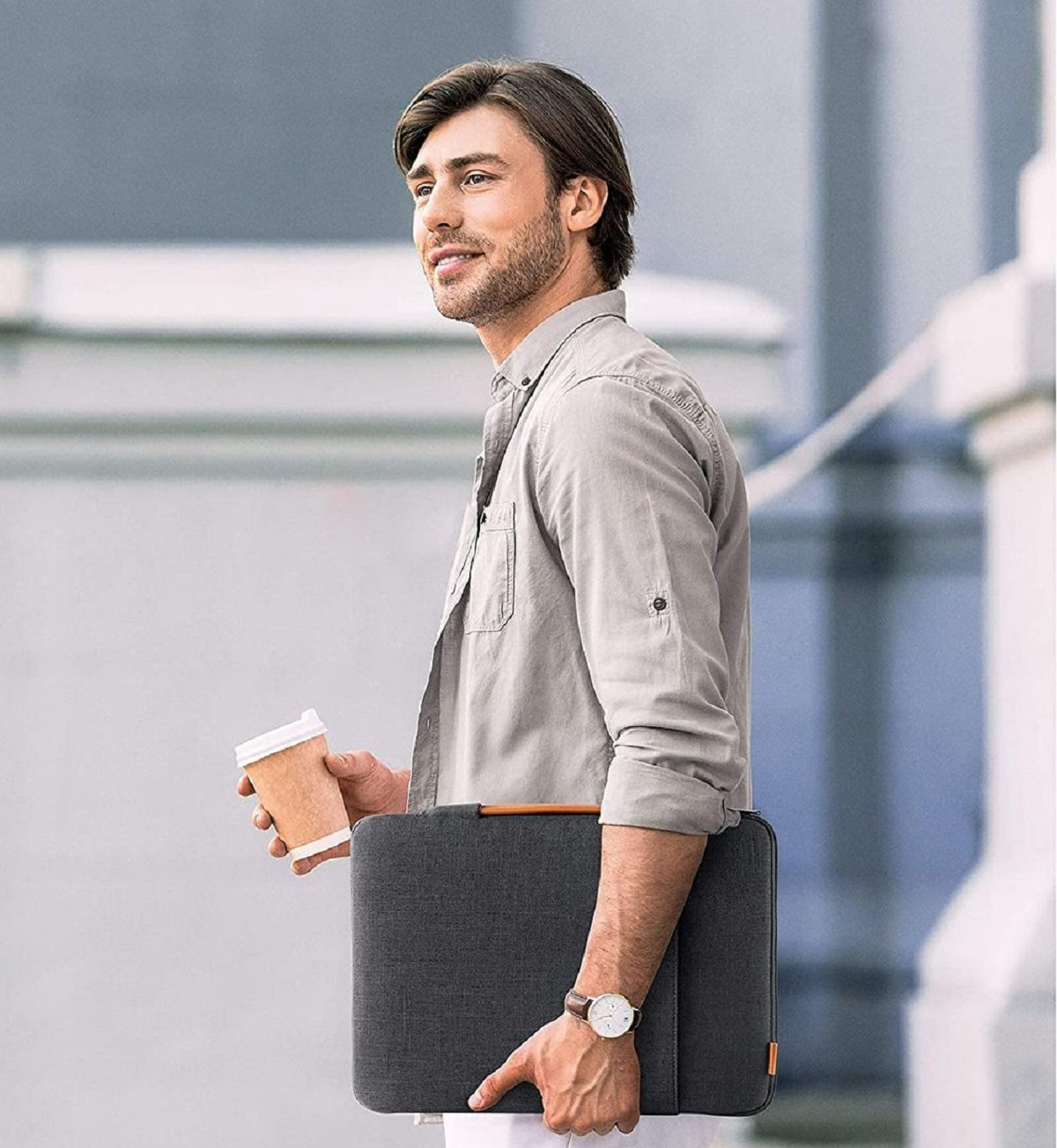 Un hombre sosteniendo una taza de café y una funda para portátil, usando un reloj, mirando hacia un lado.