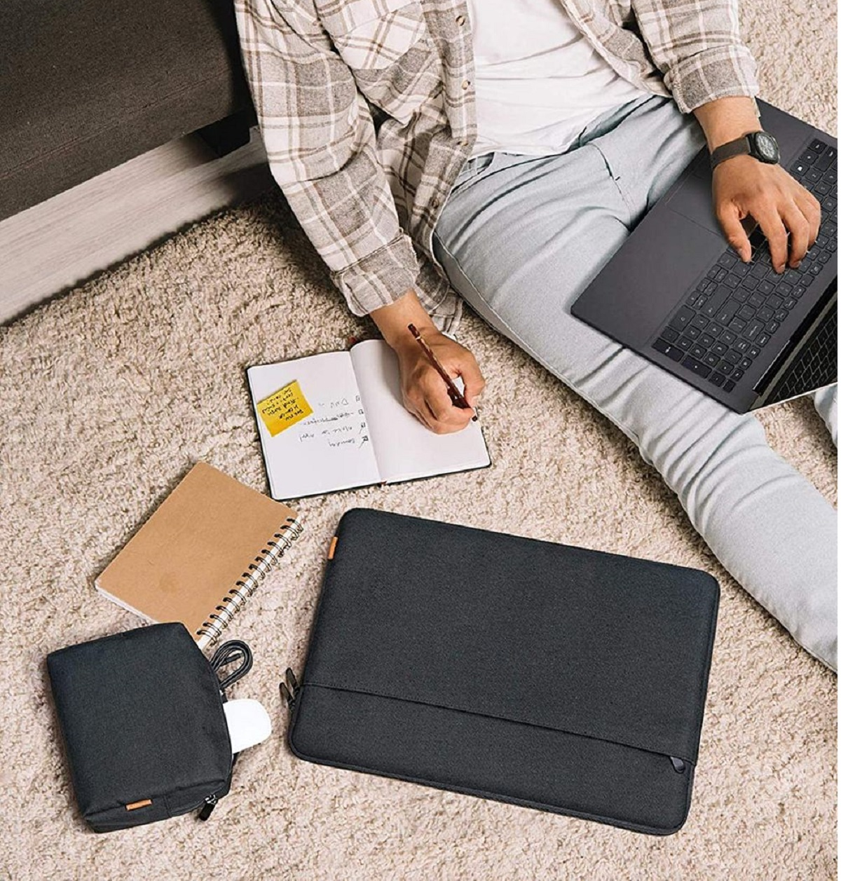 Person sitzt auf Teppich, arbeitet am Laptop, mit Notizbuch und Zubehör.