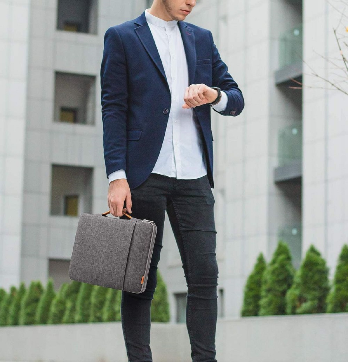 Ein Mann in einem marineblauen Blazer und schwarzer Hose schaut auf seine Uhr und hält einen grauen Aktenkoffer.